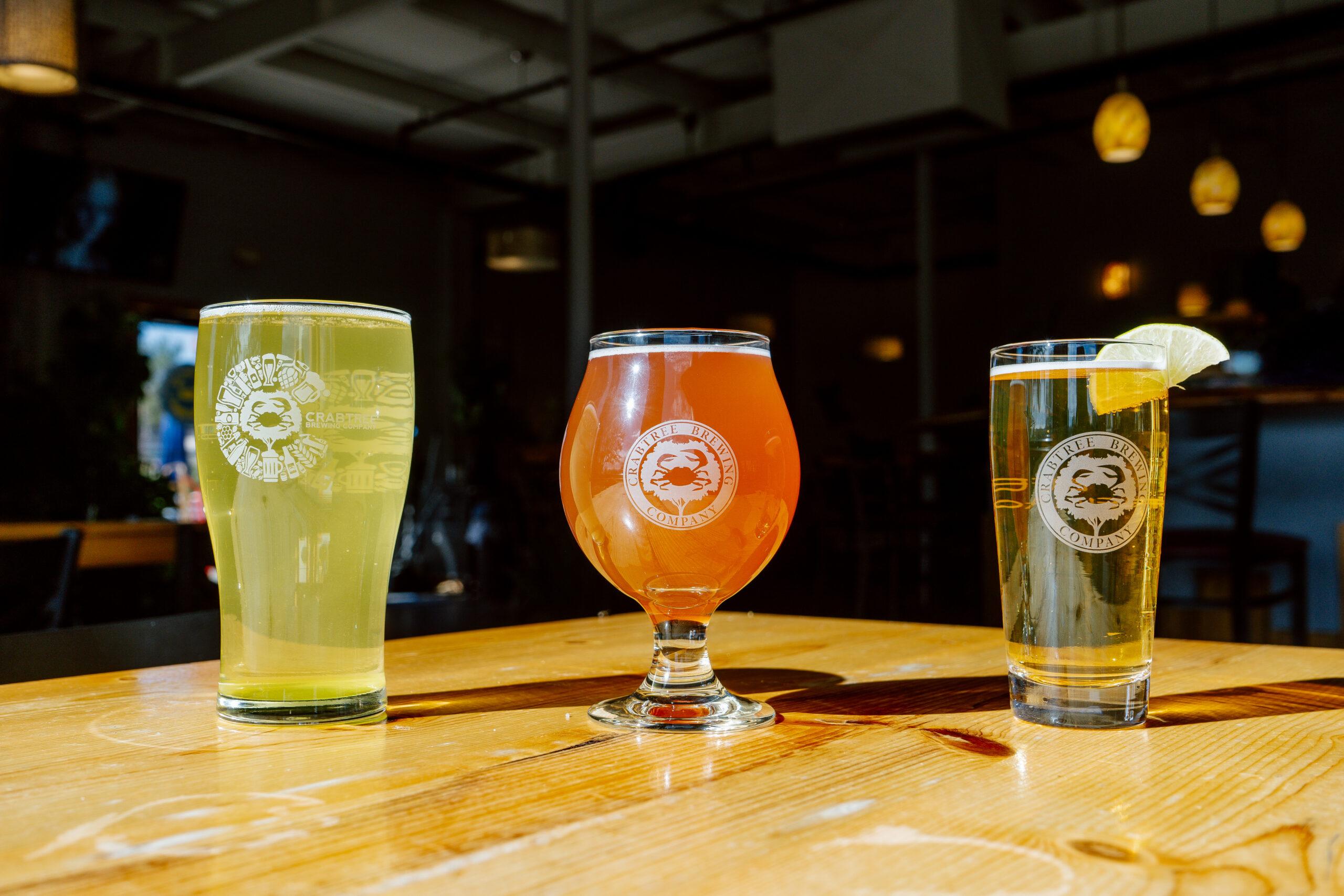 Three Crabtree beers — a pale ale, a sour, and a lager — poured in etched Crabtree glassware on a taproom table