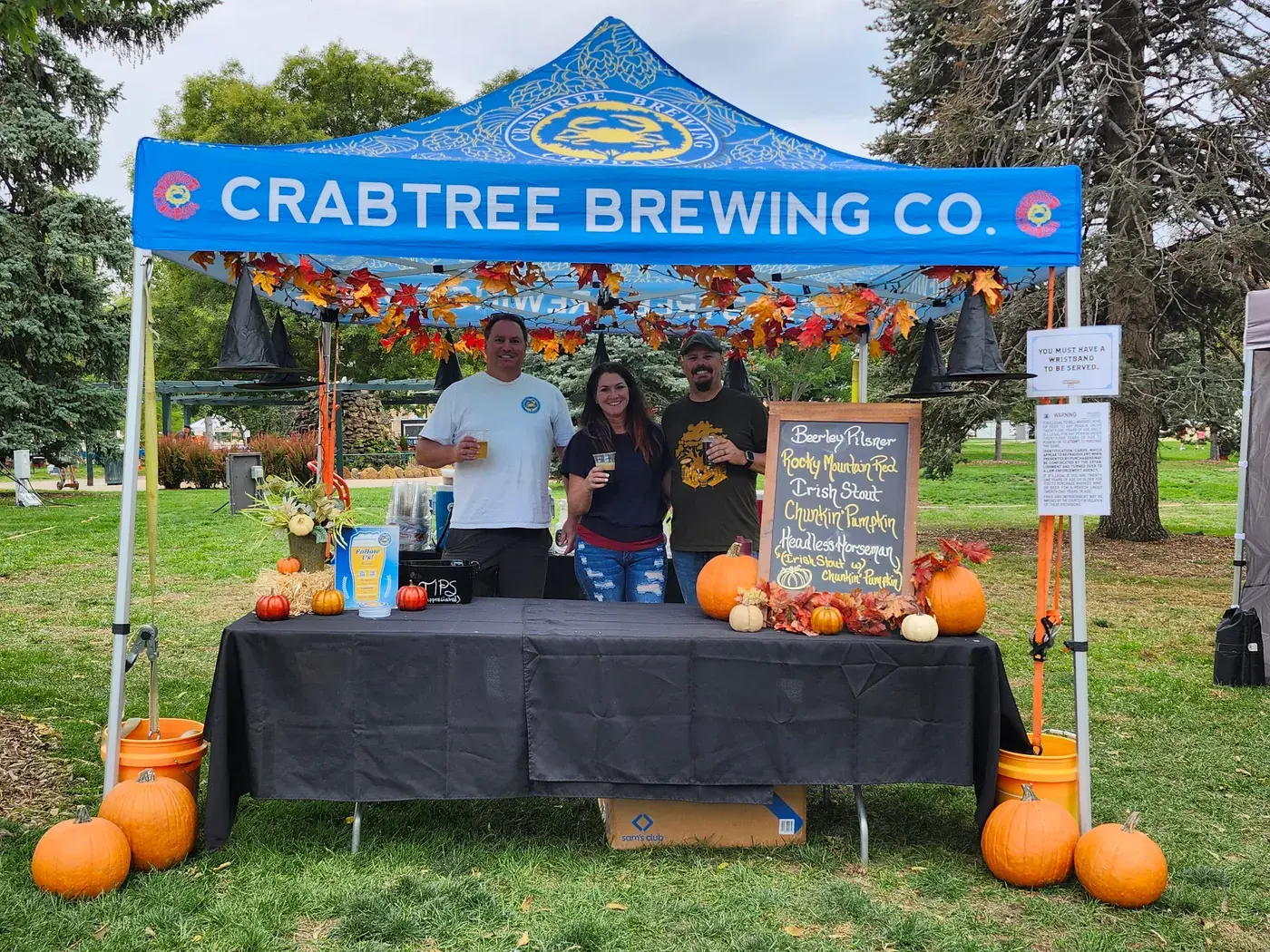 Three people raising beers at Crabtree Brewing's autumn-decorated booth at OktoBrewfest 2026